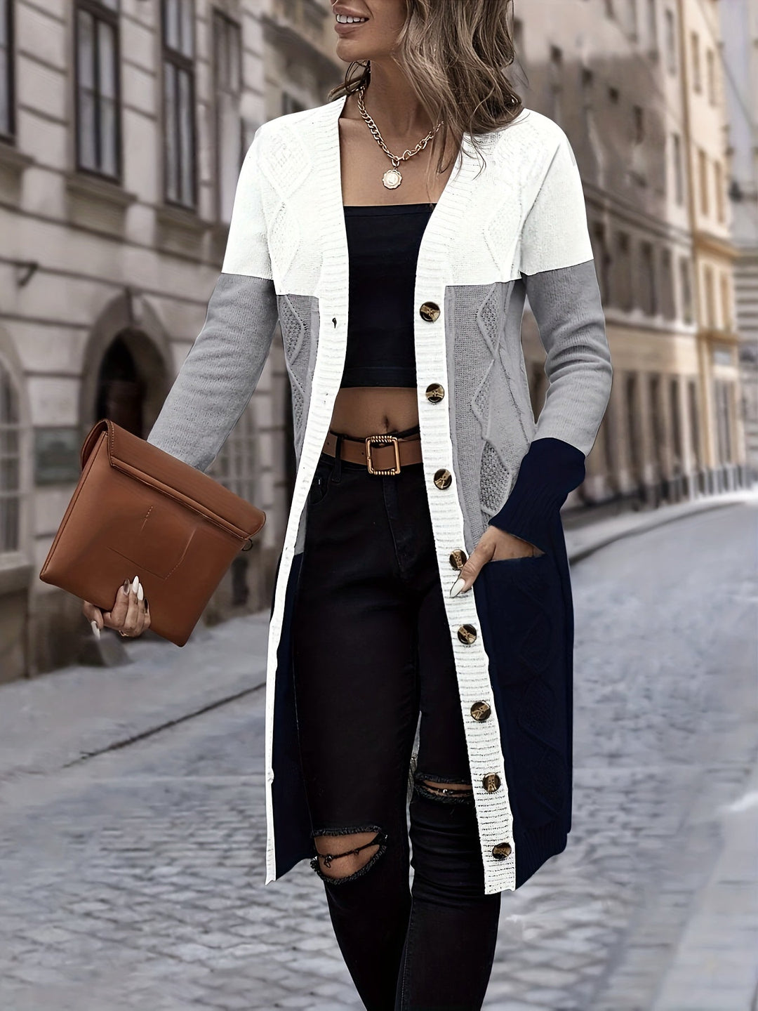 Frau in langem Strickcardigan, weiß-grau-blau, mit brauner Clutch auf Kopfsteinpflasterstraße.