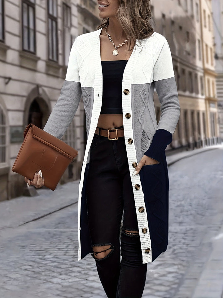 Frau in langem Strickcardigan, weiß-grau-blau, mit brauner Clutch auf Kopfsteinpflasterstraße.