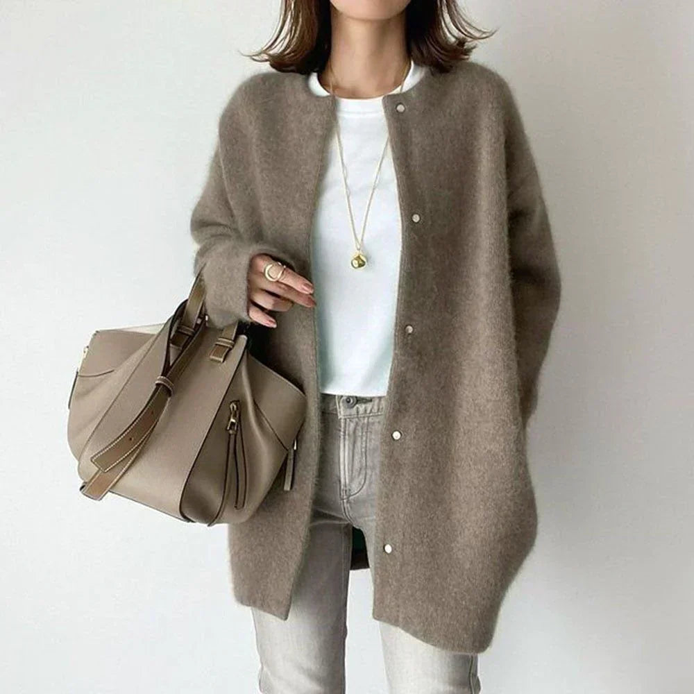 Frau in braunem Strickcardigan, weißem T-Shirt, grauer Hose, beige Handtasche, modisch.
