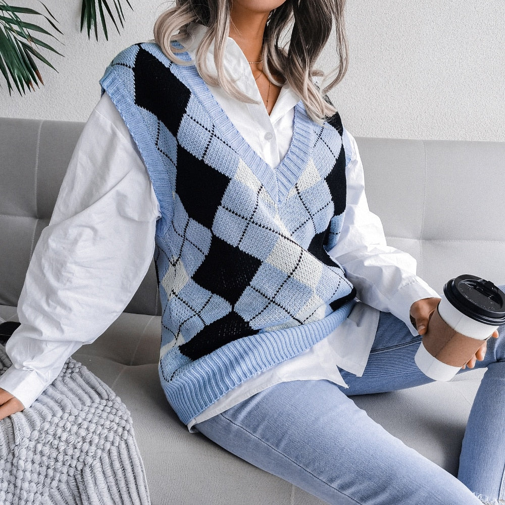 Frau in blauem Argyle-Pullover, weißem Hemd, Jeans, hält Kaffeebecher, sitzt auf Sofa.