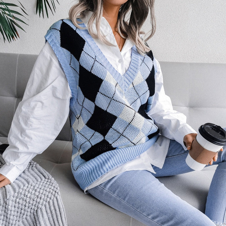 Frau in blauem Argyle-Pullover, weißem Hemd, Jeans, hält Kaffeebecher, sitzt auf Sofa.