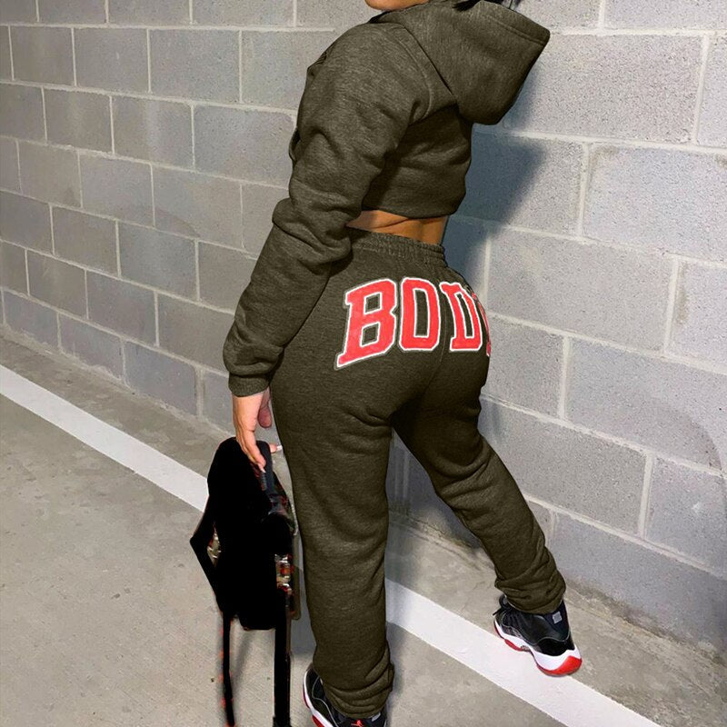 Frau in olivgrünem Jogginganzug mit "BODY"-Aufdruck, Kapuze, Turnschuhe, Betonwand.