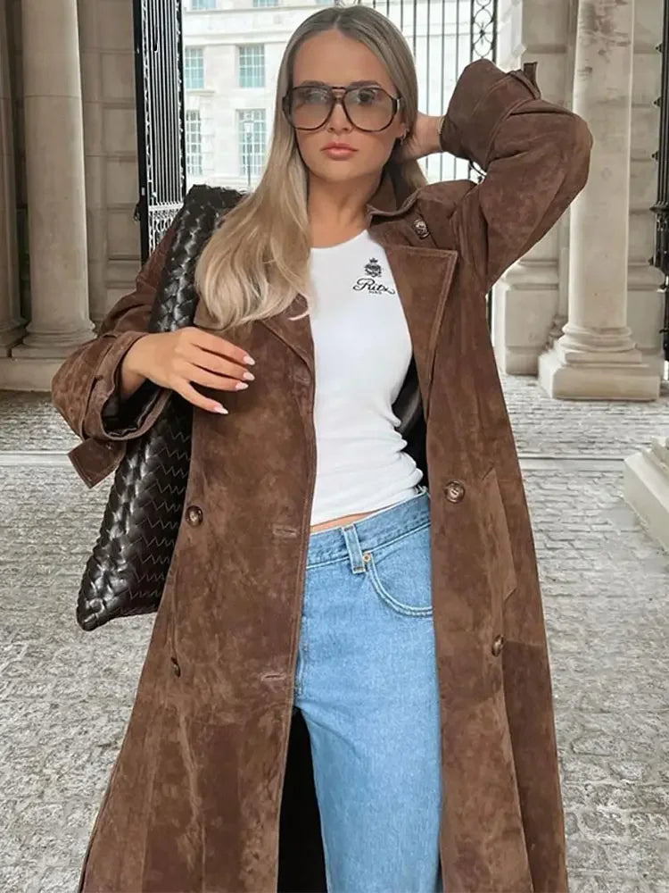 Frau in braunem Wildledermantel, weißem T-Shirt, blauer Jeans, Sonnenbrille, schwarze Tasche.