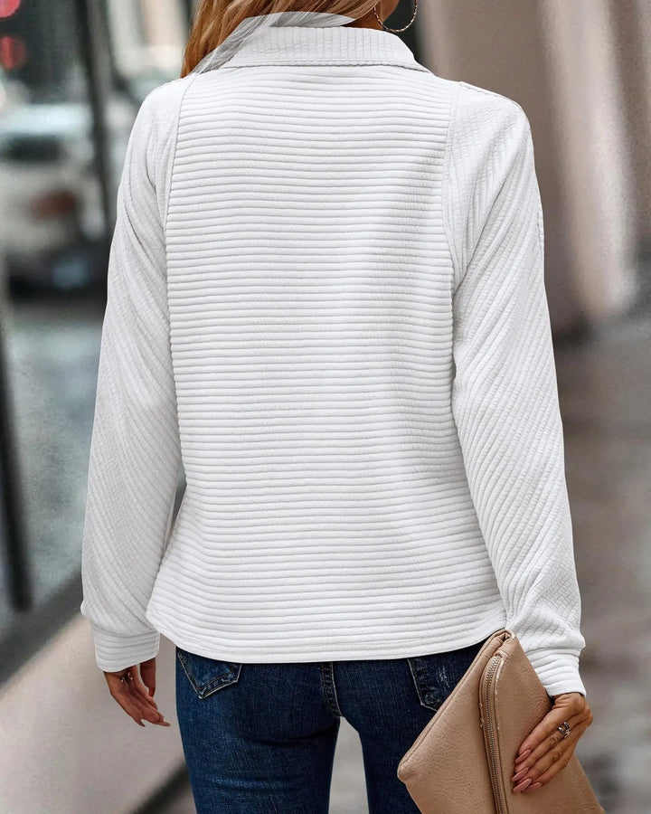 Frau in weißem Strickpullover und blauer Jeans, hält eine beige Clutch, Stadtstraße im Hintergrund.