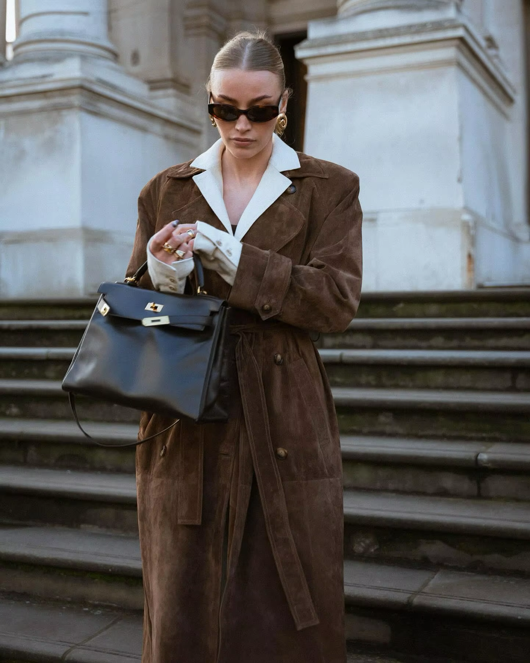 Frau in braunem Wildledermantel mit schwarzer Ledertasche und Sonnenbrille auf Treppe.
