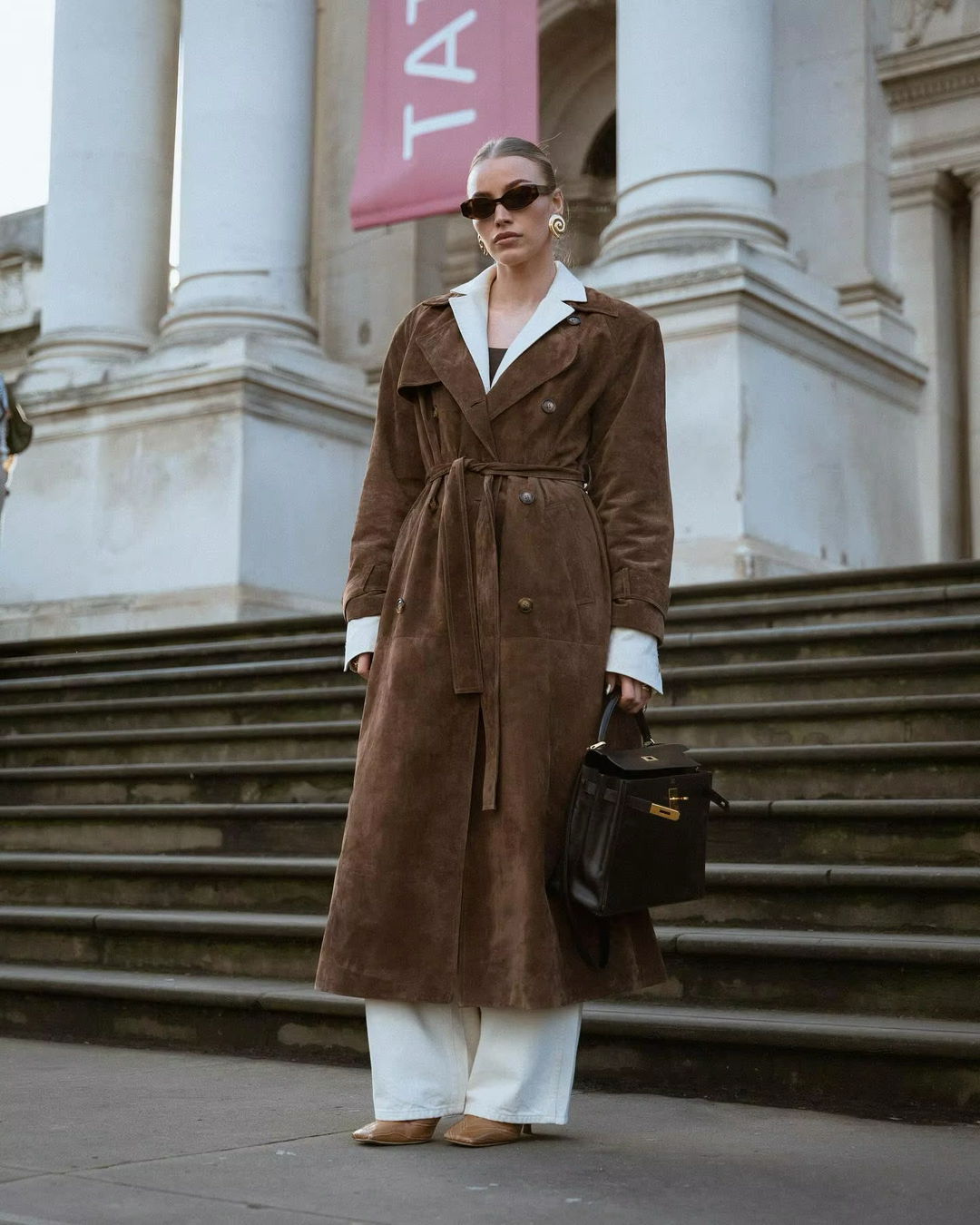 Frau in braunem Wildledermantel, weiße Hose, Sonnenbrille, schwarze Handtasche, Treppe, Stadt.