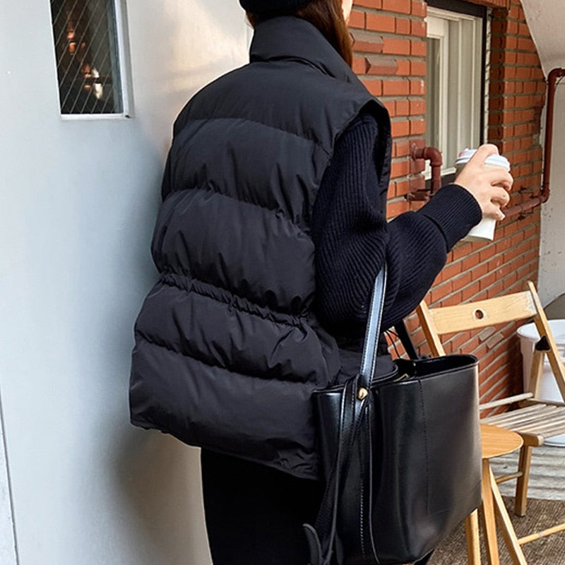 Frau in schwarzer Steppweste, hält Kaffeetasse, schwarze Tasche, Herbstmode, Ziegelwand.