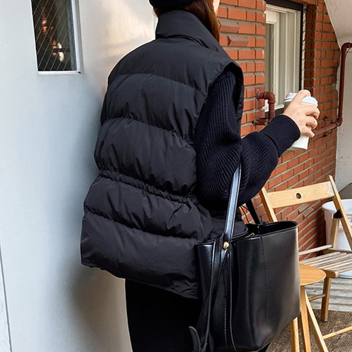 Frau in schwarzer Steppweste, hält Kaffeetasse, schwarze Tasche, Herbstmode, Ziegelwand.