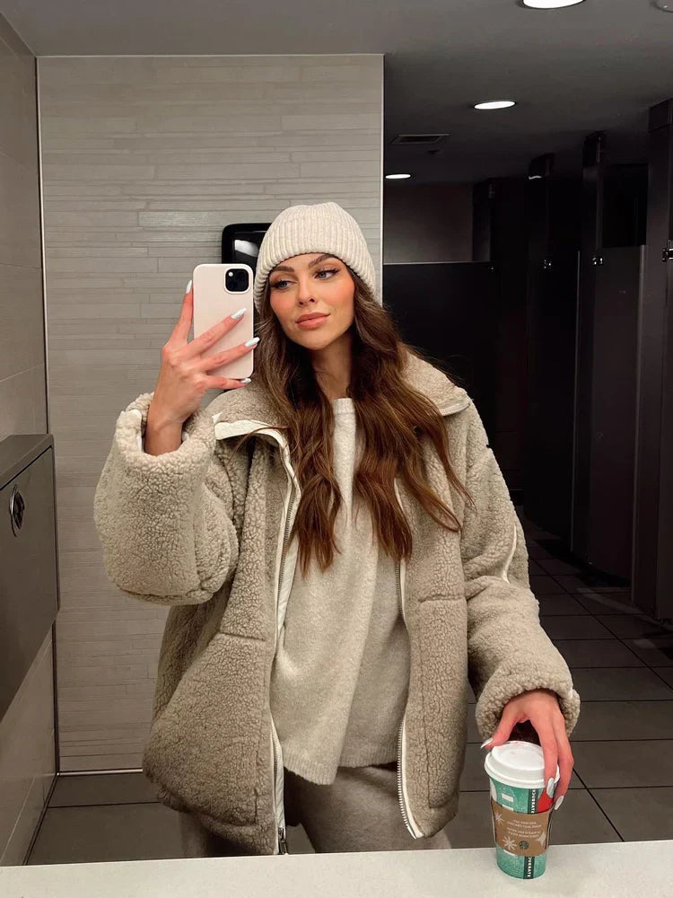 Frau im beigen Teddy-Mantel und Mütze, hält Smartphone und Kaffee, Badezimmer-Selfie.