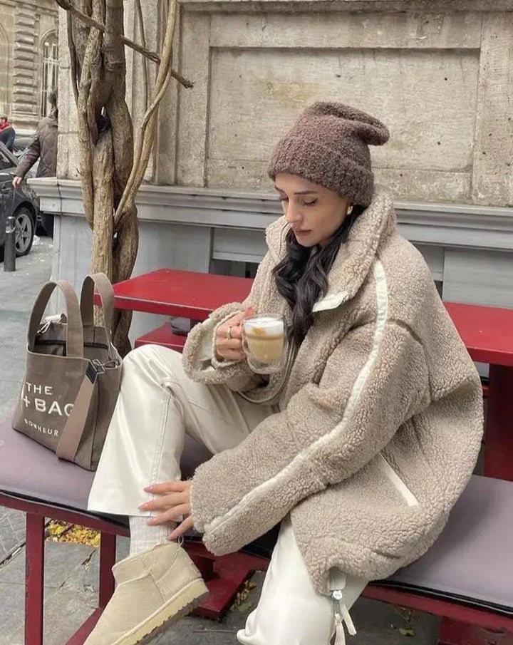 Frau in beiger Teddyjacke und Mütze, hält Kaffee, sitzt auf roter Bank, Herbstmode.