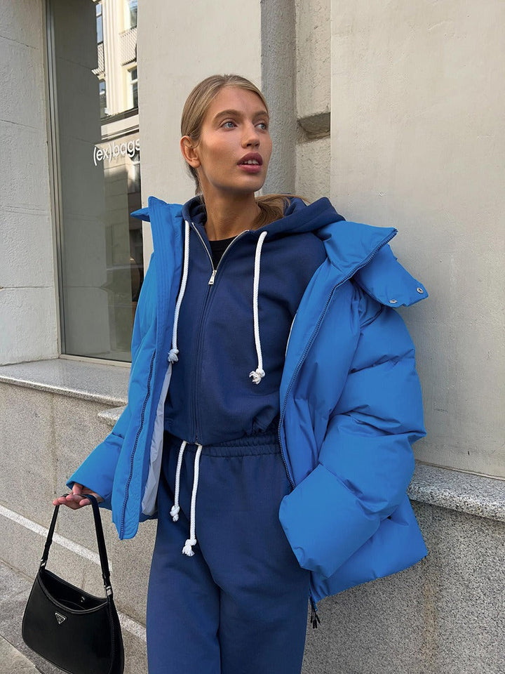 Frau in blauer Daunenjacke und Jogginganzug, hält schwarze Handtasche, Stadtumgebung.