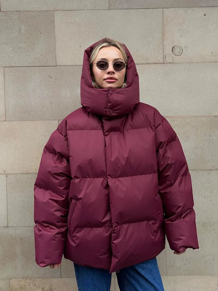 Frau in bordeauxroter Daunenjacke, Kapuze, Sonnenbrille, vor beiger Wand.