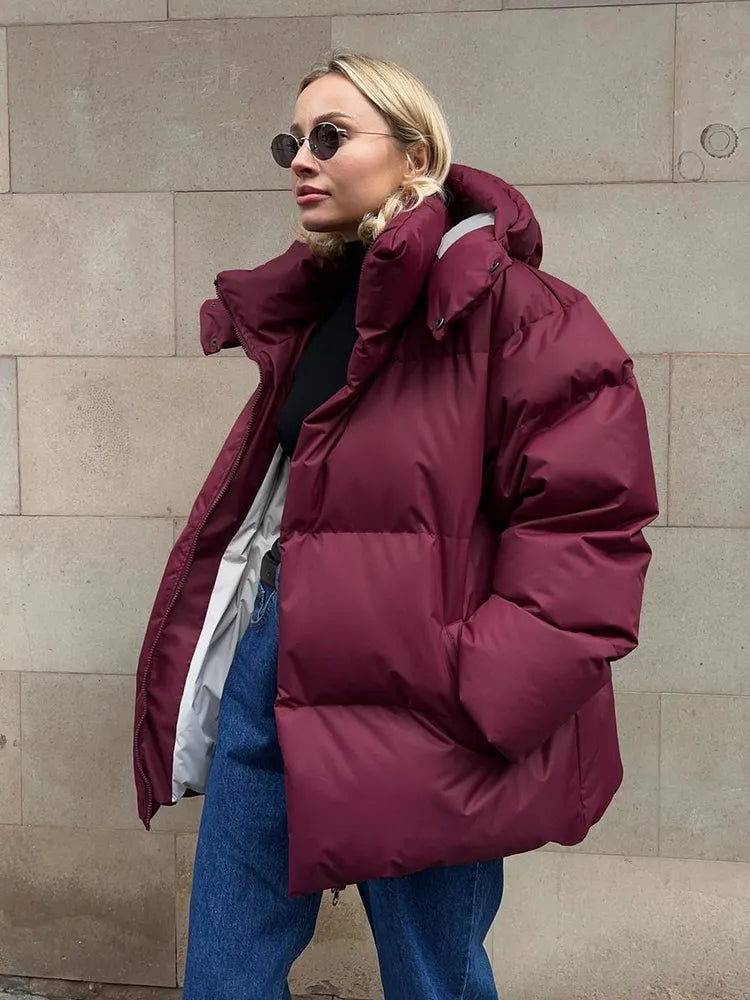 Frau in burgunderroter Daunenjacke, Sonnenbrille, Jeans, vor beiger Wand.