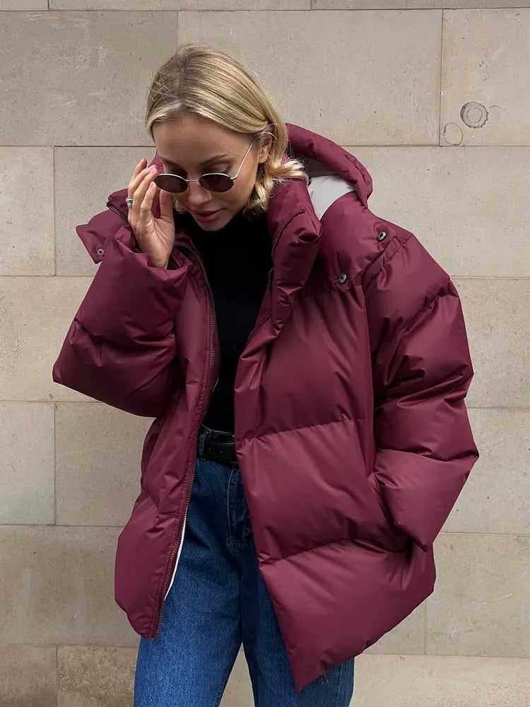 Frau in bordeauxroter Daunenjacke, Sonnenbrille, Jeans, vor beiger Wand.