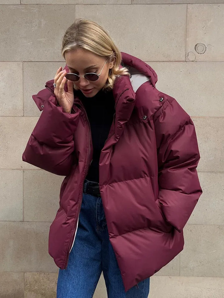 Frau in bordeauxroter Daunenjacke, Sonnenbrille, Jeans, vor beiger Wand.