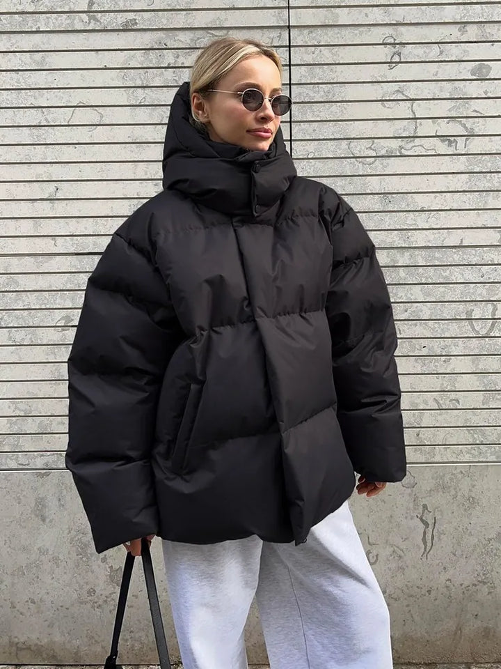 Frau in schwarzer Daunenjacke, Wintermode, stilvoll, warm, mit Sonnenbrille, vor Betonwand.