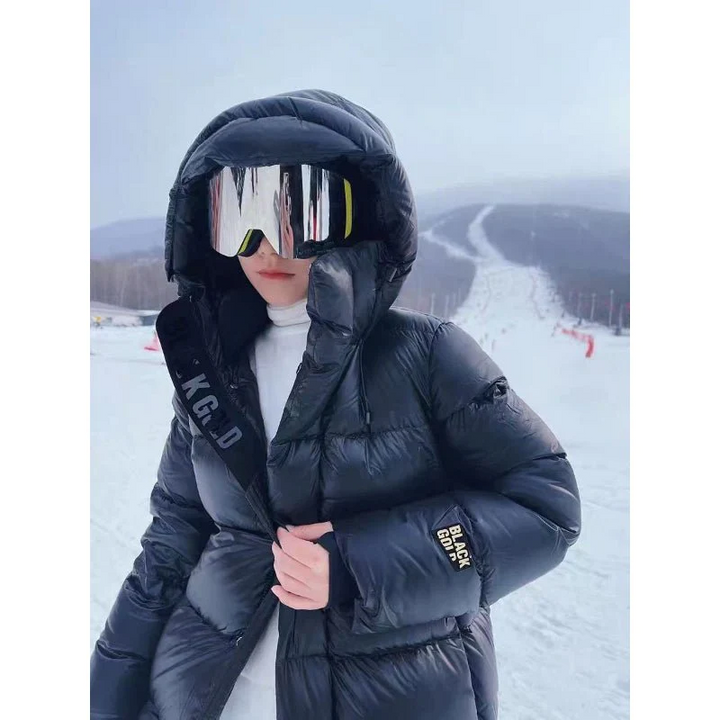 Frau in schwarzer Daunenjacke mit Kapuze und Skibrille auf verschneiter Skipiste.