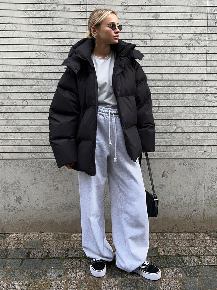 Frau in schwarzer Daunenjacke, grauem Sweatshirt, weißen Jogginghosen und Sonnenbrille.