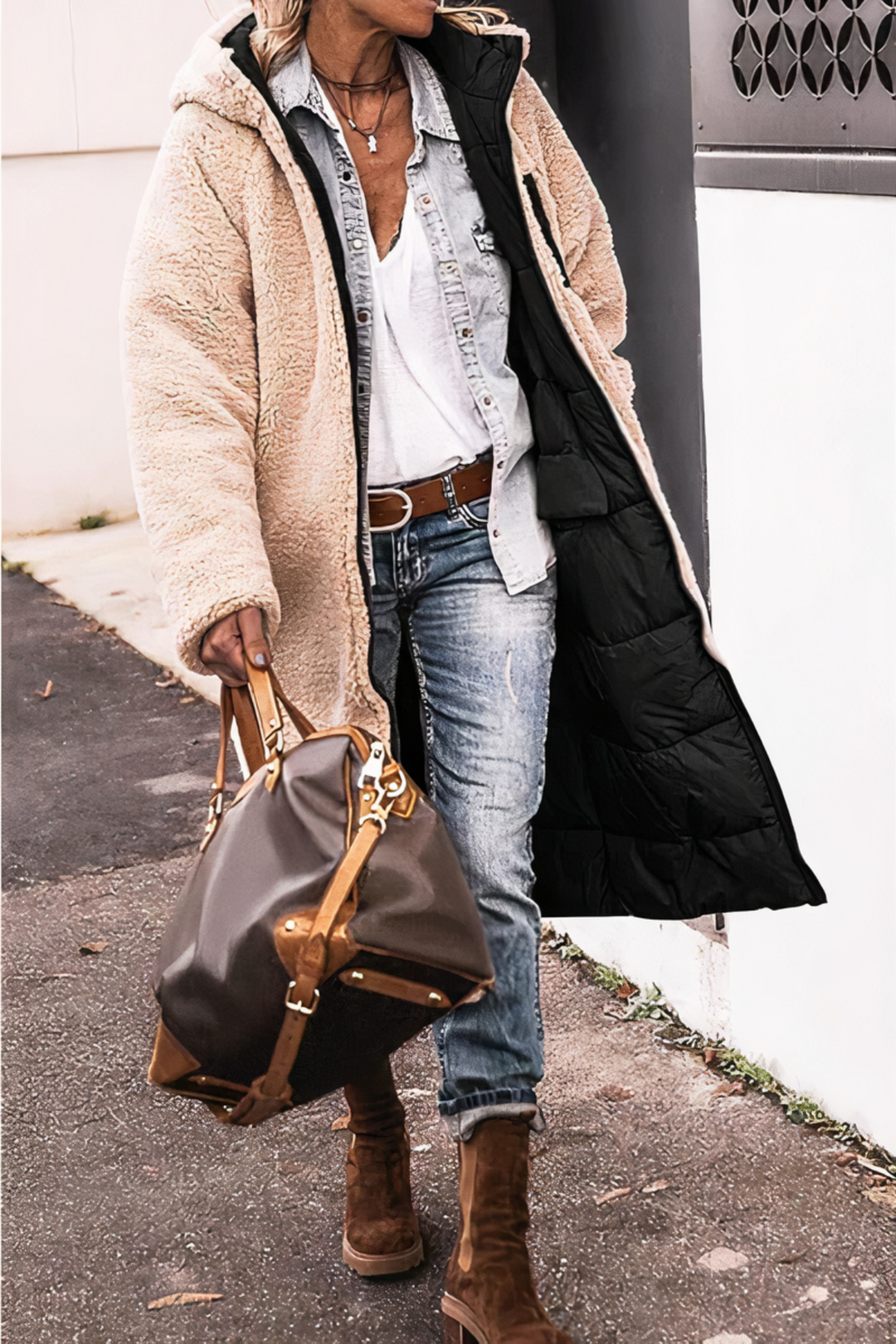 Frau in beigem Teddy-Mantel, Jeans und Stiefeln mit brauner Leder-Reisetasche auf Gehweg.