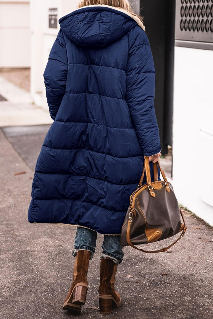 Frau in blauem Daunenmantel mit brauner Ledertasche und Stiefeln auf Straße.