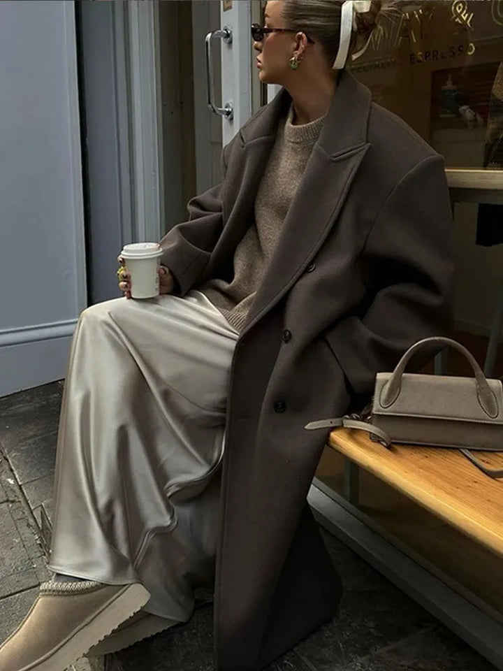 Frau in braunem Mantel, beiger Rock, hält Kaffeetasse, sitzt auf Bank, modisch, urban.