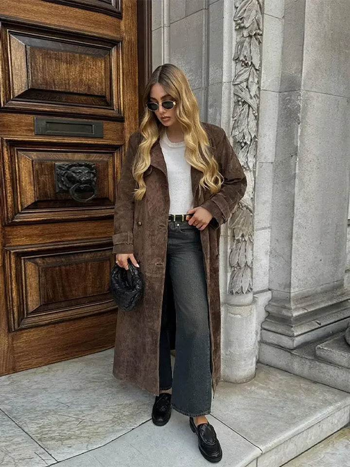 Frau in braunem Wildledermantel, weißem Shirt, schwarzer Hose, vor Holztür, modisch, elegant.