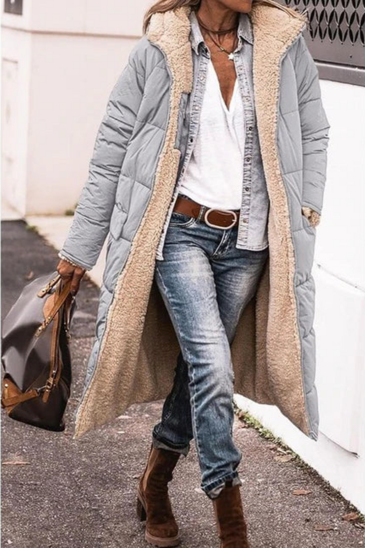 Frau in grauem Wintermantel mit Fellfutter, Jeans und braunen Stiefeln, hält braune Handtasche.