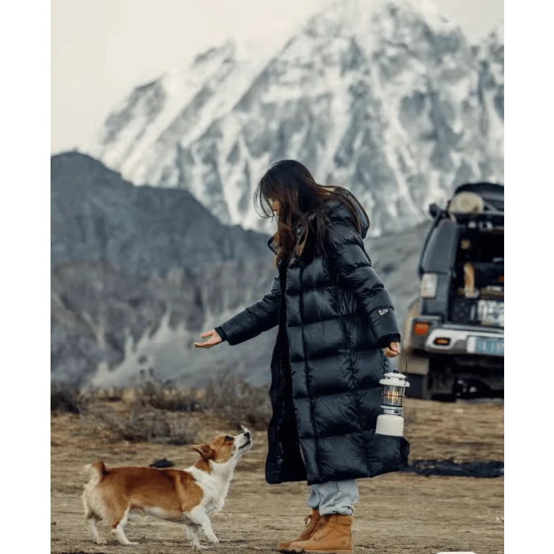Frau im langen schwarzen Daunenmantel mit Hund vor schneebedecktem Berg und Geländewagen.