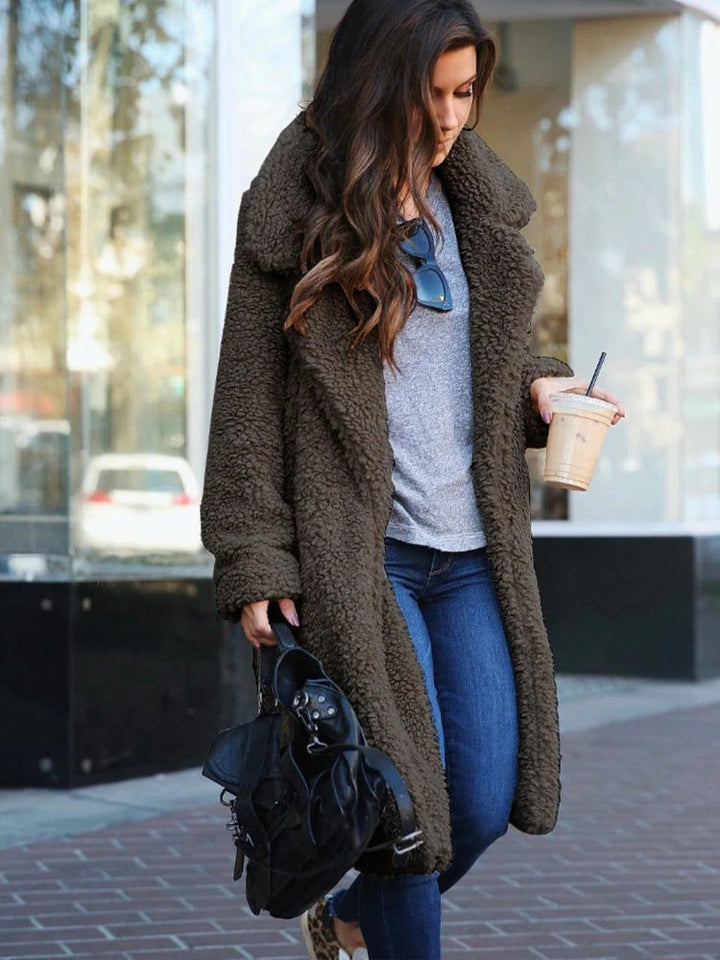 Frau in braunem Teddy-Mantel, grauem Pullover, Jeans, mit schwarzer Tasche und Eiskaffee.