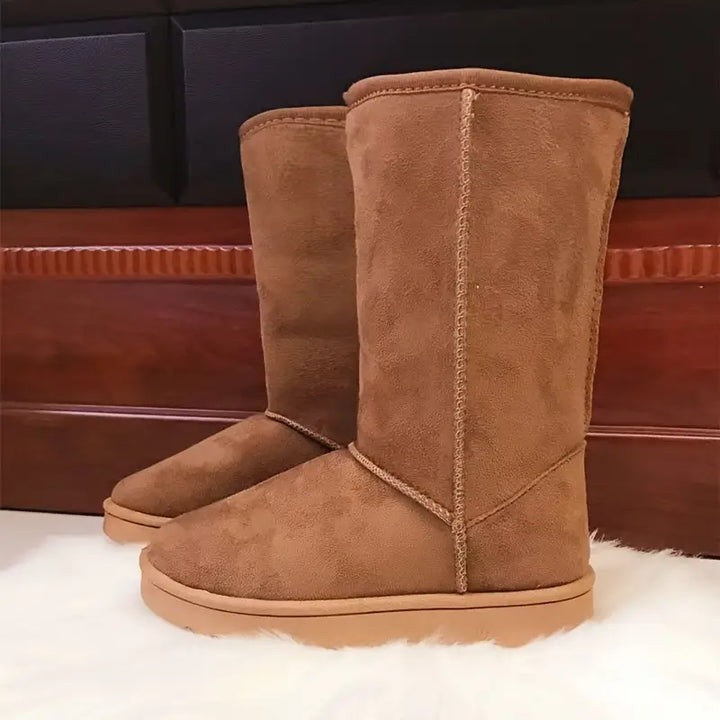 Braune Wildleder-Stiefel, kniehoch, auf weißem Teppich vor Holzwand.