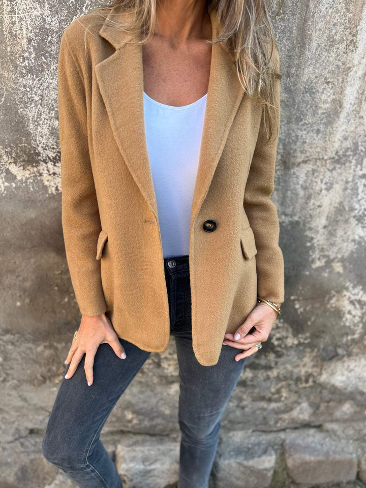Frau in braunem Wollblazer, weißem Top und grauer Jeans vor Betonwand.