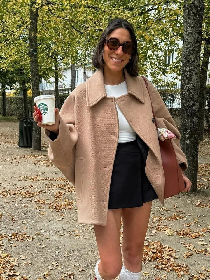 Frau im beigen Mantel mit Starbucks-Becher, schwarze Shorts, braune Tasche, Herbstpark.