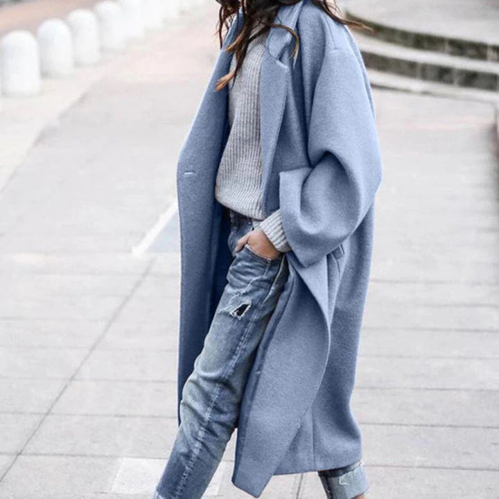 Frau in blauem Wollmantel, grauem Pullover und Jeans auf Gehweg spazierend.