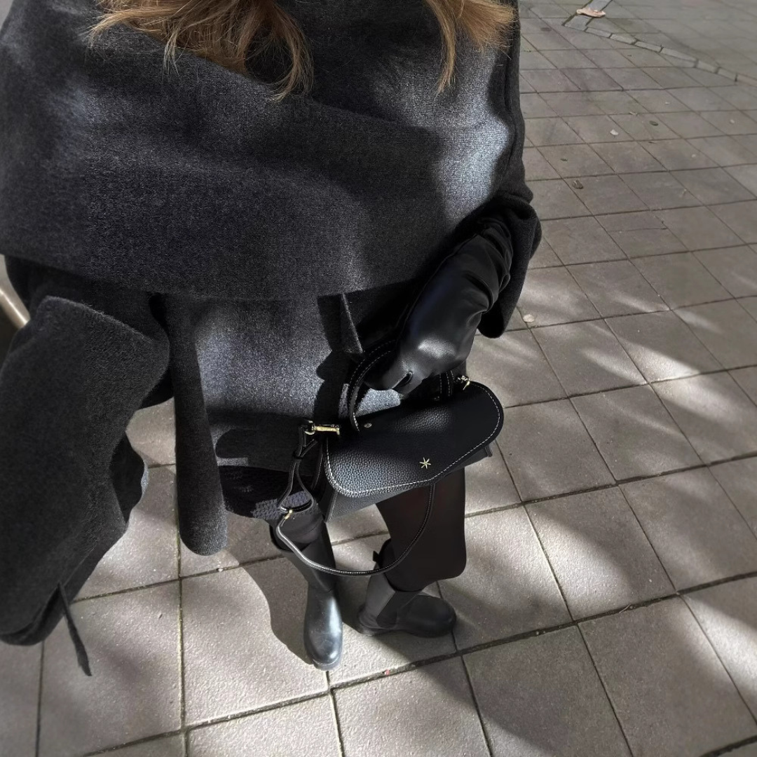 Frau in grauem Mantel mit schwarzer Lederhandtasche und Stiefeln auf gepflastertem Gehweg.