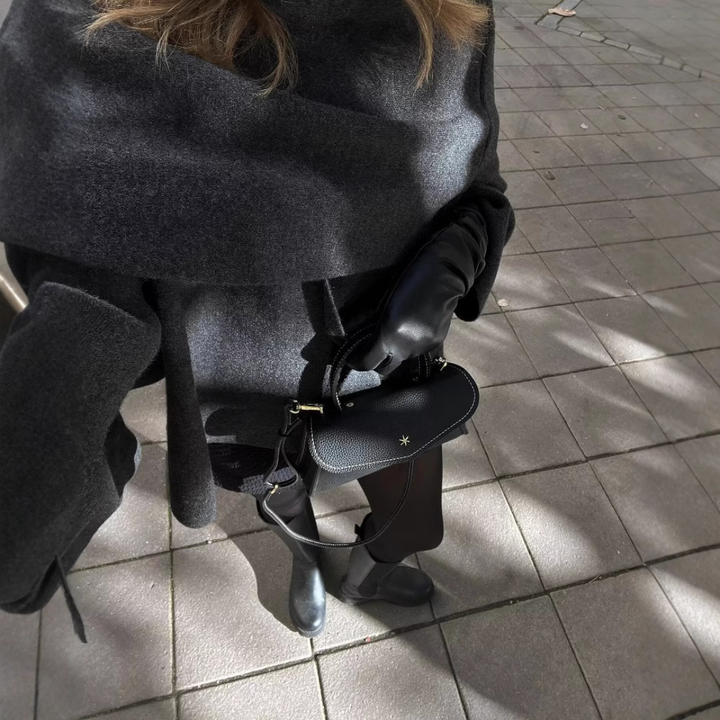 Frau in grauem Mantel mit schwarzer Lederhandtasche und Stiefeln auf gepflastertem Gehweg.
