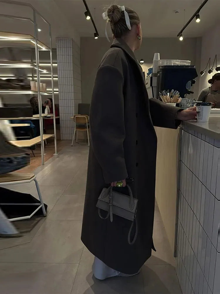 Frau im Café mit langem, dunkelgrauem Mantel und kleiner Handtasche, moderne Inneneinrichtung.