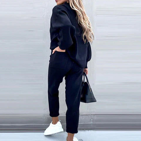 Frau in schwarzem Zweiteiler mit Rollkragen, High-Waist-Hose, weißen Sneakers, schwarze Tasche.