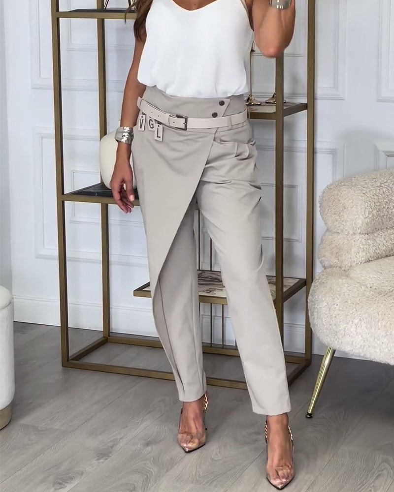 Frau in eleganter, beiger Wickelhose und weißem Top, stehend vor Regal im modernen Wohnzimmer.