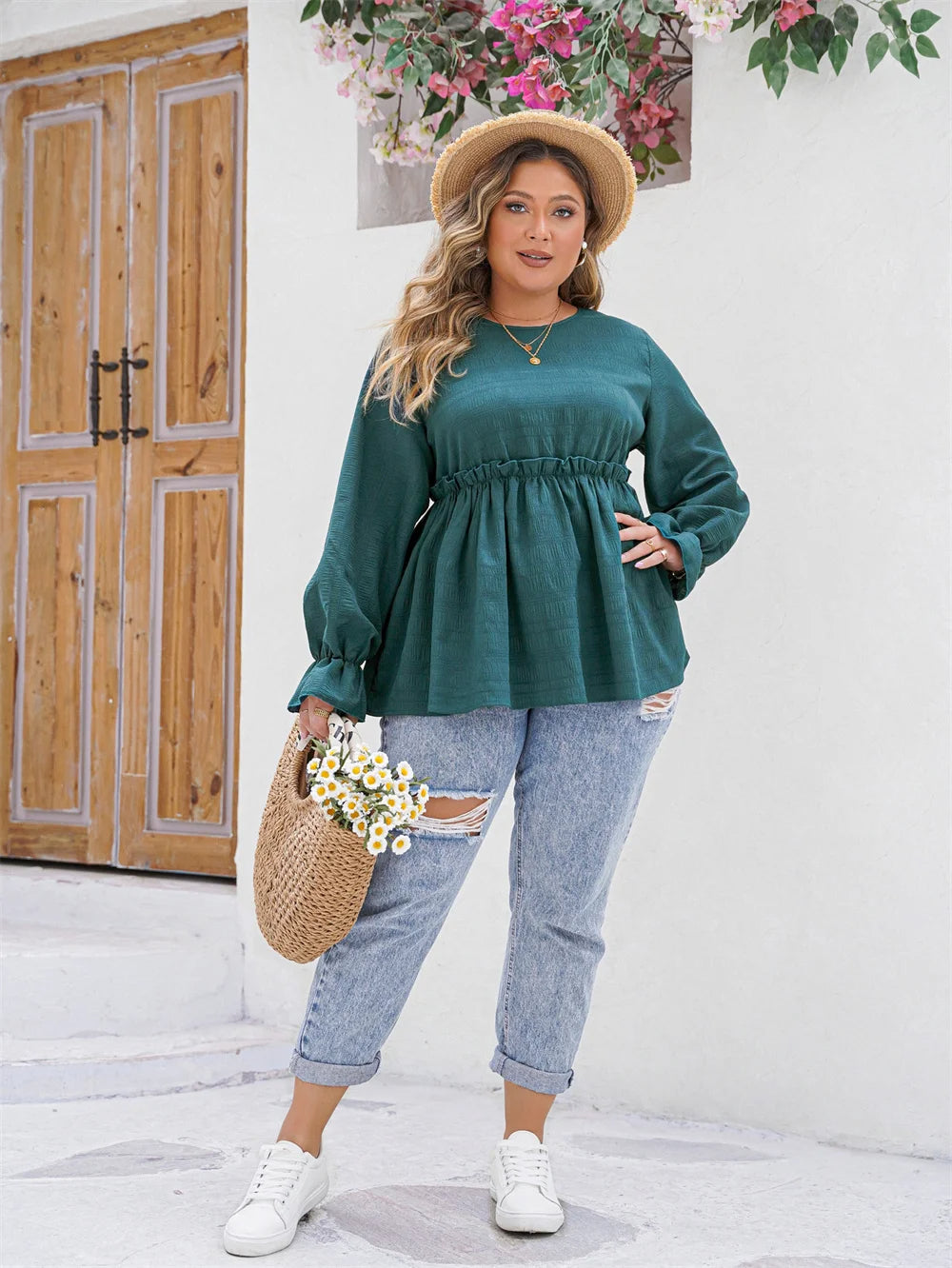 Frau in grüner Bluse und Jeans, Strohhut, Korbtasche mit Blumen, vor Holztür und Blumen.