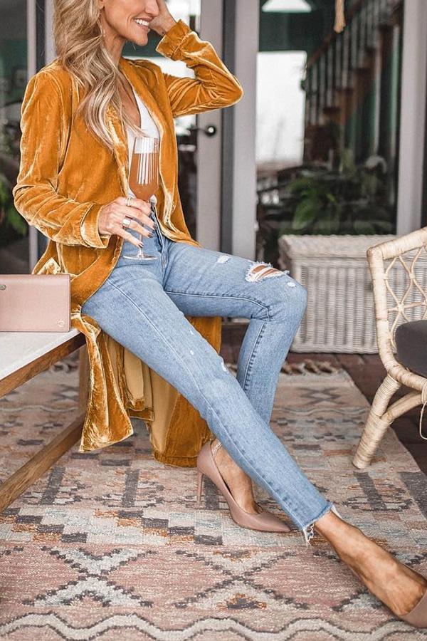Frau in gelbem Samtblazer, zerrissene Jeans, hält Sektglas, sitzt auf Teppich.
