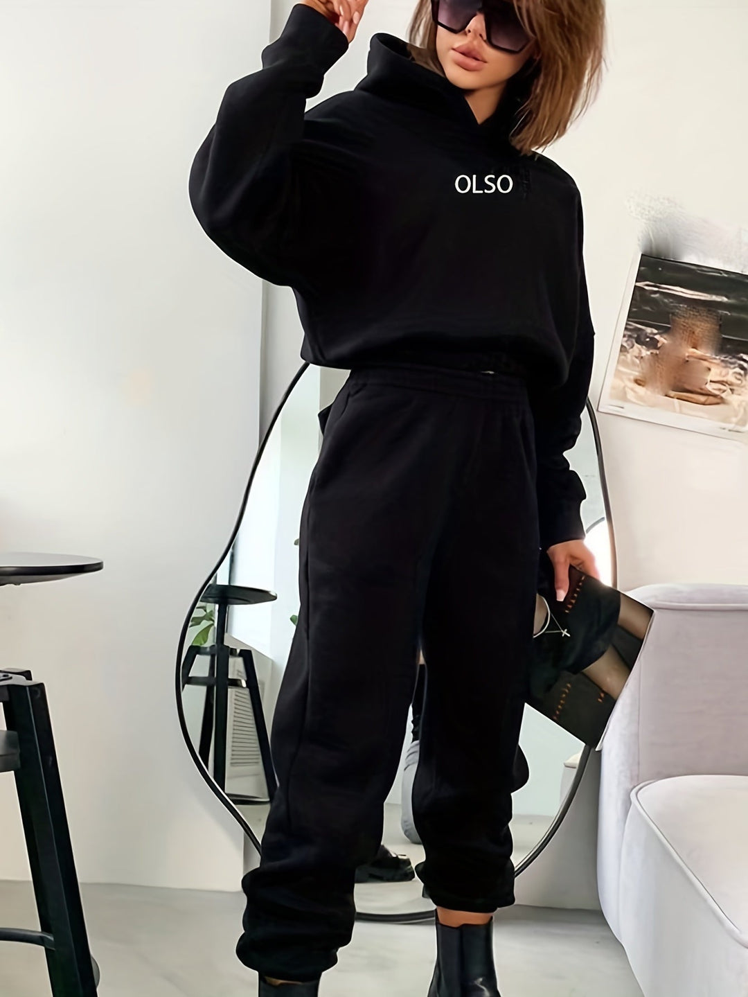 Frau in schwarzem Hoodie und Jogginghose, OLSO-Logo, modisch, lässig, Spiegel, Sonnenbrille.