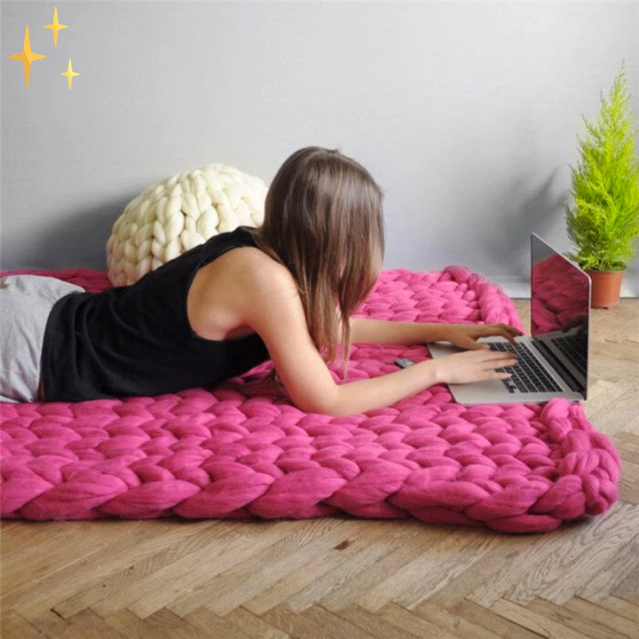 Frau auf pinker, grob gestrickter Decke, arbeitet am Laptop, gemütliches Wohnzimmer.