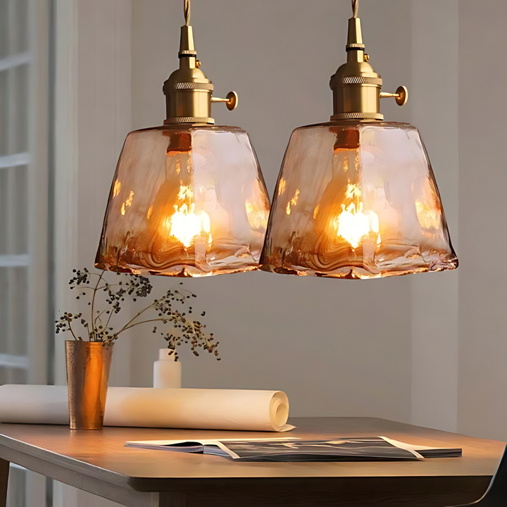 Zwei hängende, bernsteinfarbene Glaspendelleuchten über einem Tisch, warmes Licht, modernes Design.