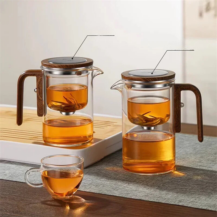 Zwei Glas-Teekannen mit Holzgriff und Teesieb, gefüllt mit Tee, auf einem Tisch.