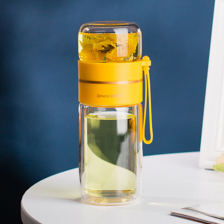 Gelbe Teeflasche mit Sieb, doppelwandig, aus Glas, auf weißem Tisch vor blauem Hintergrund.