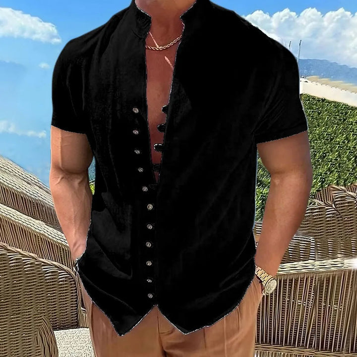Mann in schwarzem Kurzarmhemd mit Knopfleiste, braune Hose, Strandhintergrund.