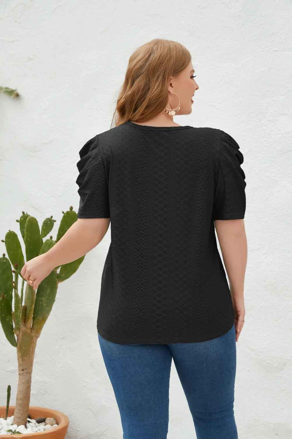 Frau in schwarzem T-Shirt mit Puffärmeln, blaue Jeans, vor weißer Wand, neben Kaktus.