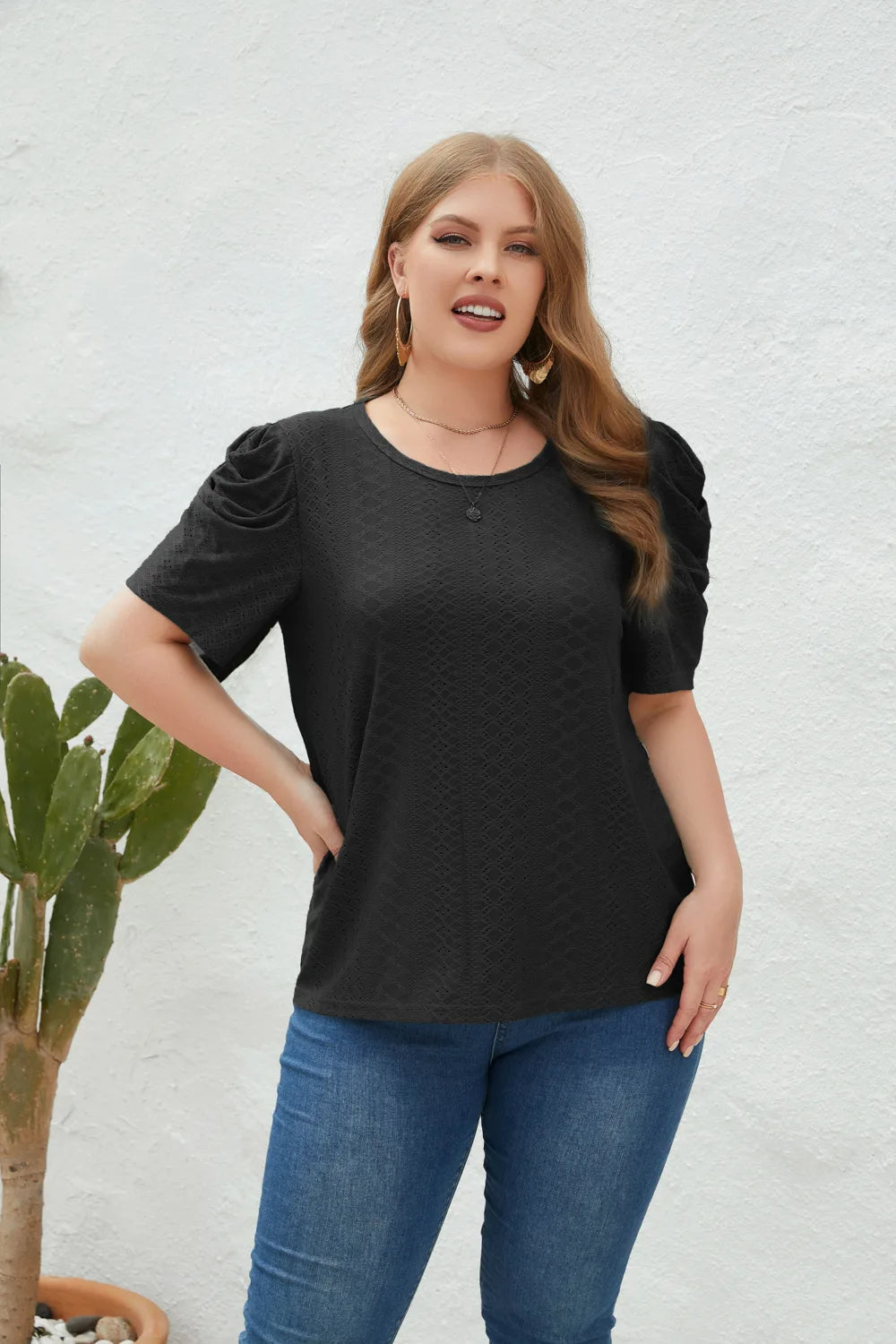 Frau in schwarzem T-Shirt mit Puffärmeln, blaue Jeans, vor weißer Wand, neben Kaktus.