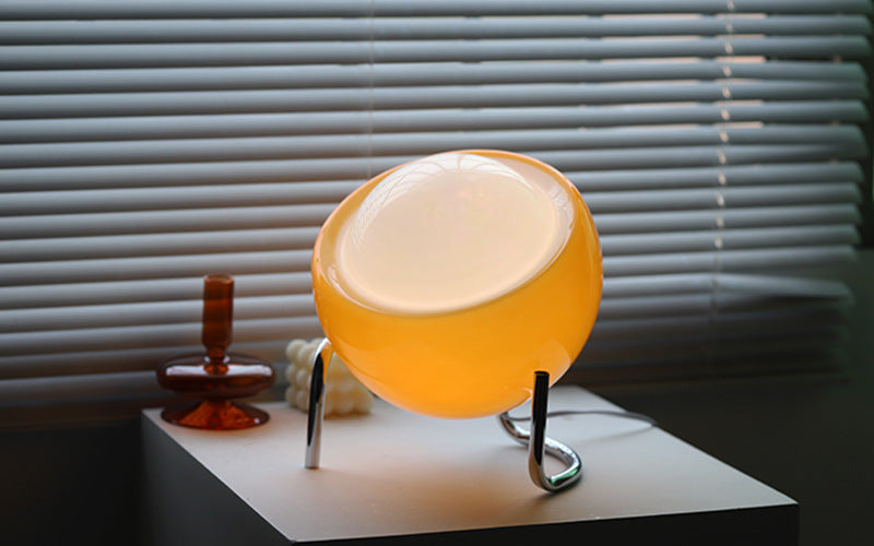 Gelbe Tischlampe aus Glas auf weißem Tisch vor Jalousien, modernes Design, Wohnzimmerbeleuchtung.