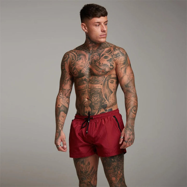 Mann mit Tattoos trägt rote Sportshorts, muskulös, Fitnessmode, Studioaufnahme.